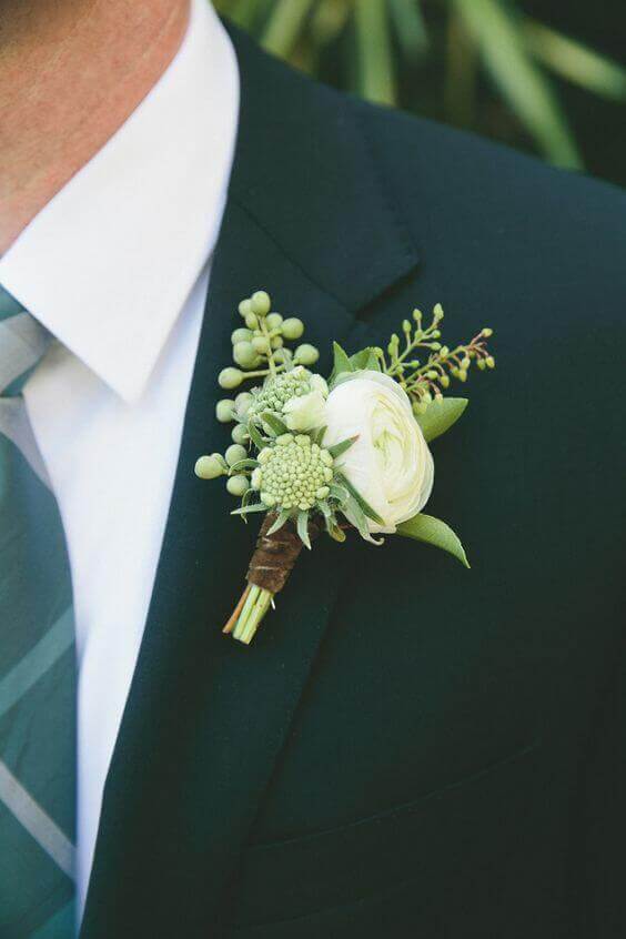 38 wedding accessories list - ideas to transform your big day into magic 12 Close-up of a wedding boutonniere featuring a white ranunculus and green berries, elegantly pinned to a dark green suit lapel.