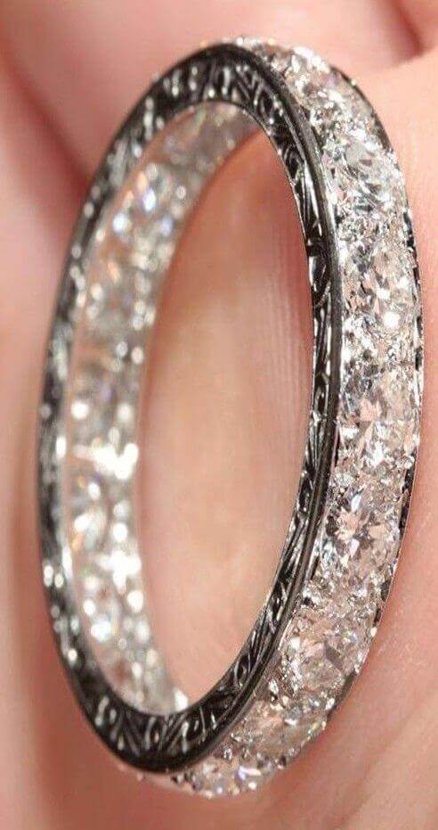 Macro close-up of a diamond eternity wedding band in platinum or white gold, intricately engraved and sparkling, held delicately between fingertips.