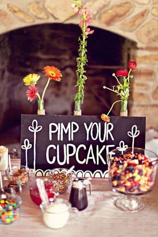 34 budget wedding ideas for a creative affordable wedding 13 Rustic wedding cupcake bar featuring a chalkboard sign "pimp your cupcake," colorful candy toppings, syrups, and vibrant flowers by a stone fireplace.