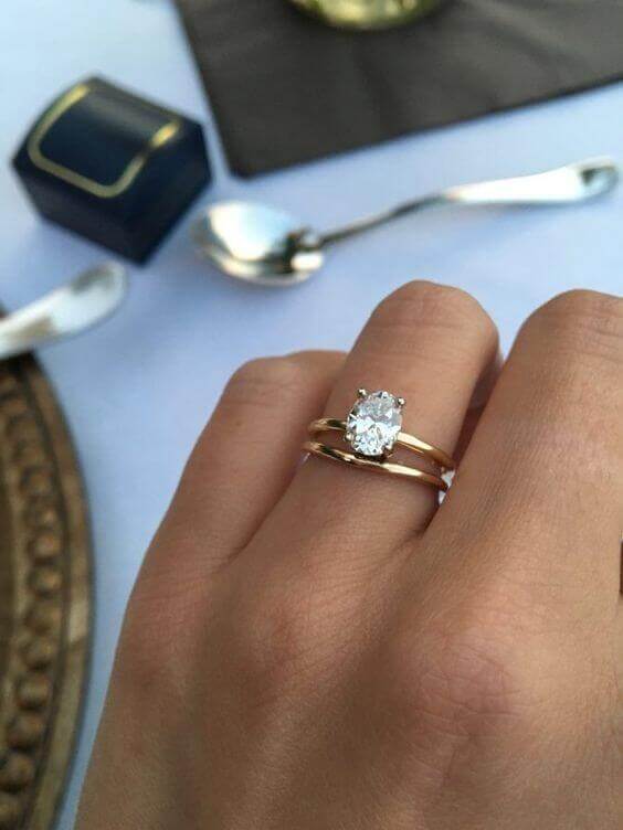 Hand adorned with an oval diamond solitaire and slim yellow-gold wedding band, set against a blurred backdrop of a navy ring box and elegant tableware.