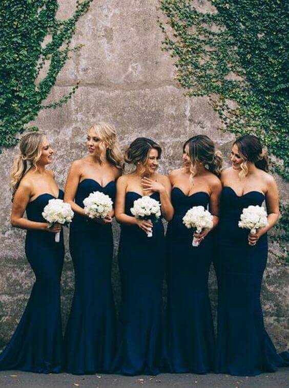 31 steps wedding planning checklist 13 Five bridesmaids in navy strapless gowns hold white rose bouquets, smiling against an ivy-covered stone wall, embodying wedding elegance.