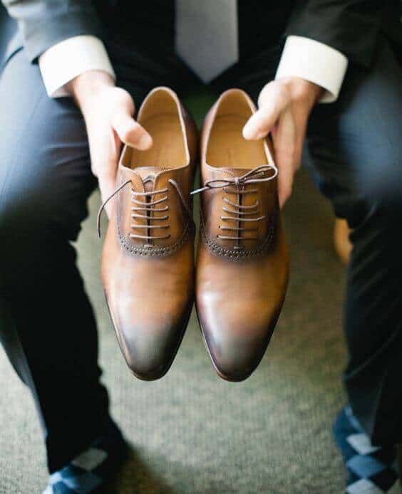 38 wedding accessories list - ideas to transform your big day into magic 14 Groom in dark suit holding polished cognac-brown oxford shoes, preparing for the ceremony in a softly lit getting-ready suite.