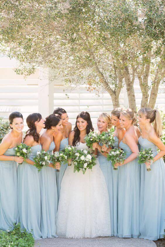 Bride in an ivory lace gown with bridesmaids in dusty-blue dresses, all holding green-and-white bouquets under a sunlit garden tree.