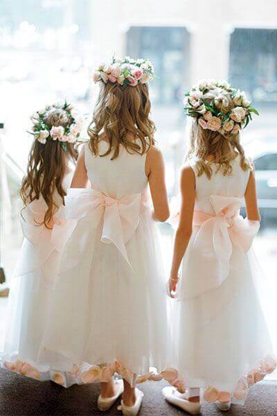 43 cute and perfect flower girl dresses that make every little princess shine 15 Three flower girls in white tulle dresses with blush organza bows stand at a wedding venue's entrance, backlit by natural daylight.