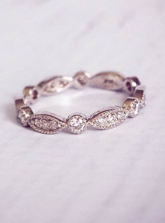 Vintage-inspired diamond eternity wedding band in white gold, featuring milgrain details, marquise pavé stations, and a soft bokeh background.