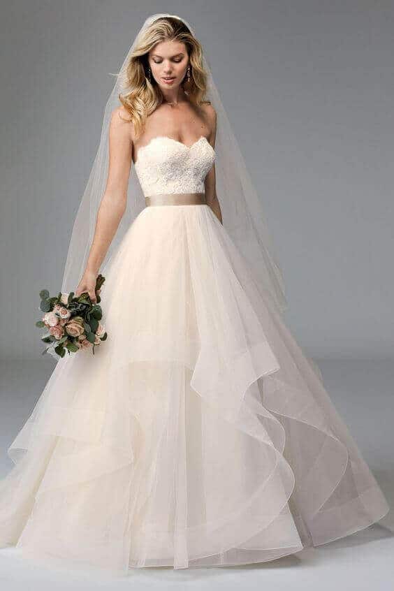 31 steps wedding planning checklist 15 Bride in a strapless sweetheart ivory gown with layered tulle skirt, champagne belt, and blush rose bouquet, against a gray backdrop.