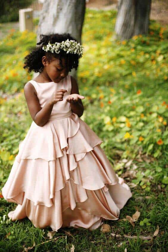 43 cute and perfect flower girl dresses that make every little princess shine 16 A flower girl in a blush pink tiered dress stands in a garden, holding a petal, surrounded by greenery and yellow-orange blooms.