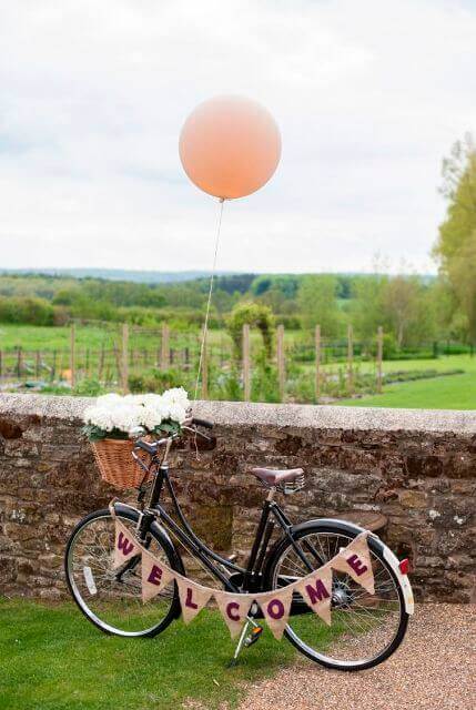 34 budget wedding ideas for a creative affordable wedding 17 Rustic wedding vignette with a black vintage bicycle, peach balloon, and welcome bunting against a stone wall in a lush countryside.