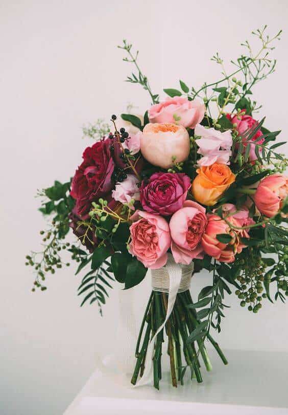 38 wedding accessories list - ideas to transform your big day into magic 17 Hand-tied bridal bouquet featuring blush, peach, coral, and burgundy flowers, airy greenery, and ivory ribbon, displayed on a white pedestal.