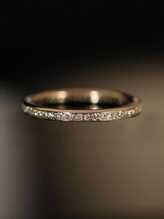 A macro shot of a slender eternity-style wedding band, featuring micro-pavé diamonds on a polished white-metal shank, glowing against a dark backdrop.
