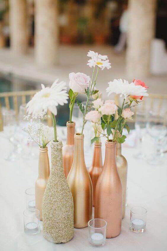 34 budget wedding ideas for a creative affordable wedding 19 Cluster of rose gold, copper, and glittered bottle vases with white and blush flowers, surrounded by clear votive candles on an ivory wedding table.