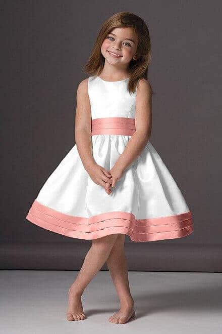43 cute and perfect flower girl dresses that make every little princess shine 20 White flower girl dress with a blush pink sash and hem bands, knee-length a-line skirt, sleeveless bodice, child smiling in studio setting.