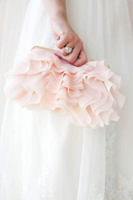 38 wedding accessories list - ideas to transform your big day into magic 21 Bride's hand delicately holds a blush ruffle clutch against an ivory wedding dress. The scene captures soft textures and a sparkling ring.