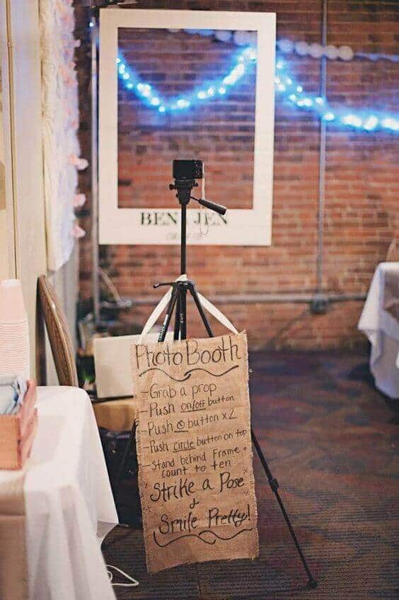 34 budget wedding ideas for a creative affordable wedding 22 Rustic wedding photo booth with a compact camera on a tripod, oversized white frame, and blue string lights, set against a brick wall.