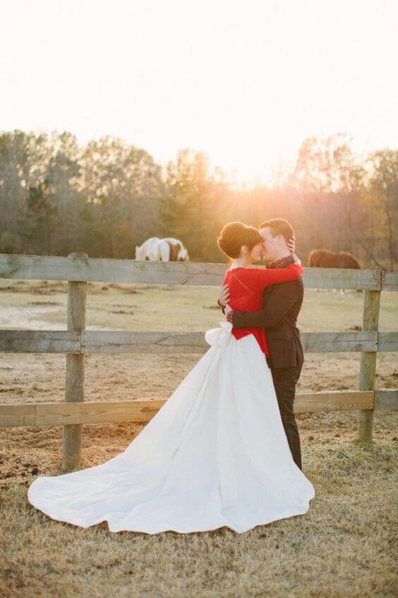 38 wedding accessories list - ideas to transform your big day into magic 22 Bride in a white ballgown with a long train and a bold red wrap embraces a groom in a dark suit at sunset, with horses grazing nearby.