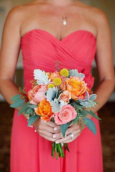 Coral-clad bridesmaid holding a vibrant hand-tied bouquet of peach and orange roses, yellow billy balls, and eucalyptus—a stunning bridesmaid dress idea.
