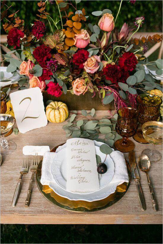 31 autumn wedding flowers ensembles perfect for your special day 22 Autumn wedding tablescape featuring a lush burgundy and blush floral centerpiece, gold flatware, and amber goblets on a wooden farm table.