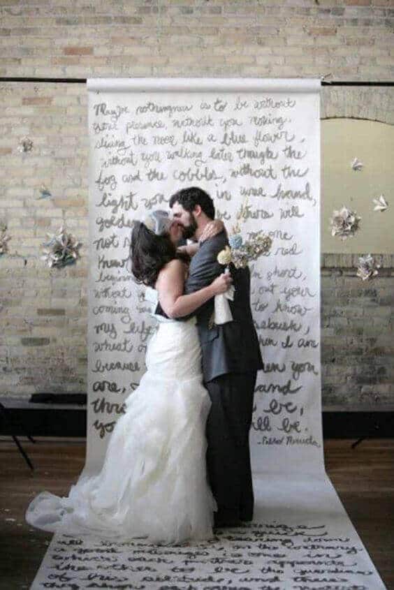 34 budget wedding ideas for a creative affordable wedding 24 Bride in ivory gown and groom in charcoal suit kiss before a calligraphy backdrop, creating an intimate budget wedding scene.
