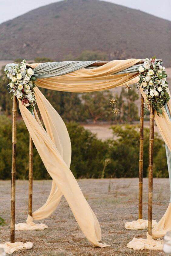 34 budget wedding ideas for a creative affordable wedding 25 Bohemian bamboo wedding arch draped in champagne and sage chiffon, adorned with white florals and gold stars, set in a meadow with a mountain backdrop.