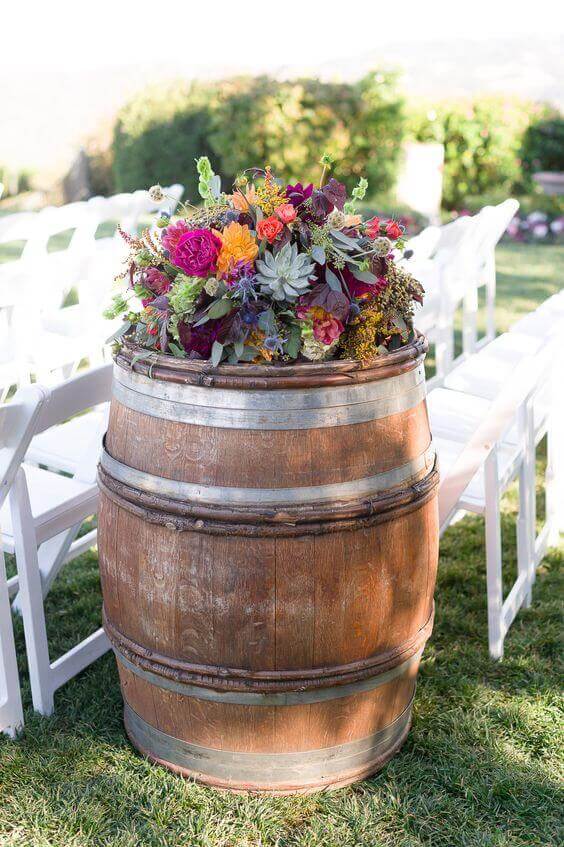 31 autumn wedding flowers ensembles perfect for your special day 24 A weathered wine barrel adorned with a lush floral arrangement of dahlias, roses, and succulents serves as an aisle marker for a rustic autumn wedding. Rows of white chairs line the sunlit grass, framed by soft-focus hedges and hills.