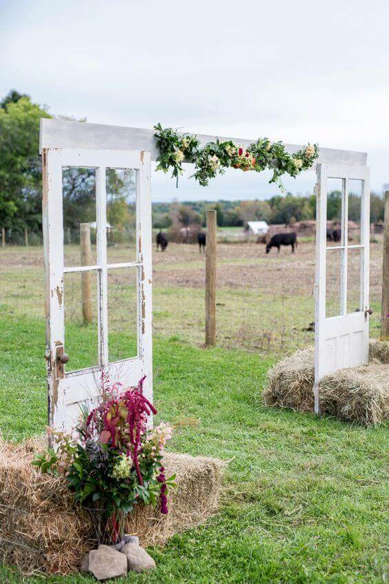 34 budget wedding ideas for a creative affordable wedding 26 Rustic wedding entrance featuring two distressed white vintage doors, a floral garland, and hay bales, set in a pastoral backdrop.