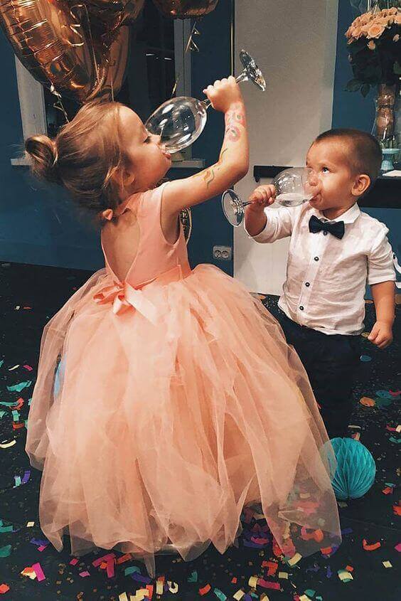43 cute and perfect flower girl dresses that make every little princess shine 26 Two children at a flower girl reception mimic toasting with glasses; girl in peach tulle dress with satin bow, boy in white shirt and bow tie, surrounded by confetti and balloons.