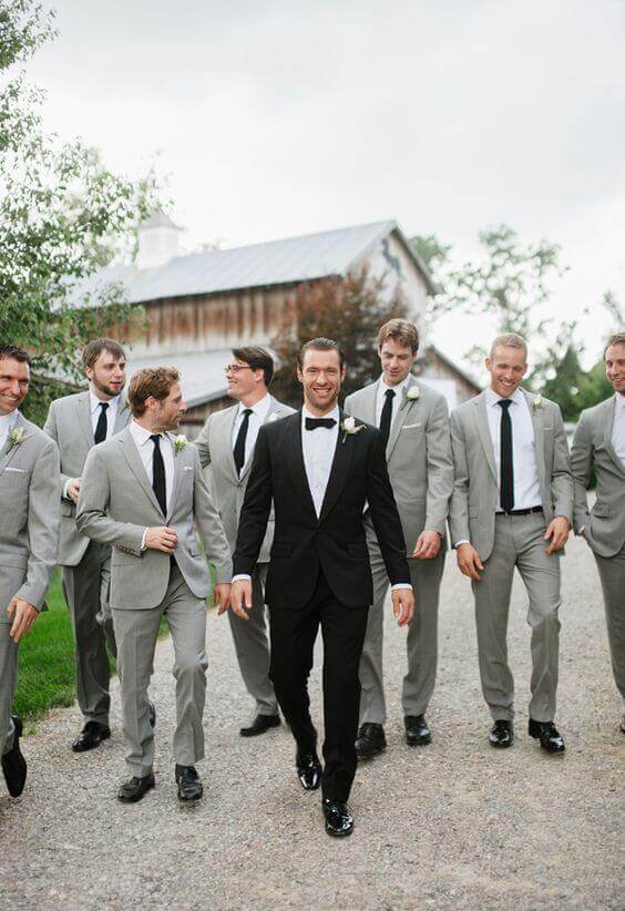 31 steps wedding planning checklist 26 {{post_main_keyword}}: groom in a black tuxedo walks with groomsmen in light gray suits down a gravel lane beside a rustic barn.