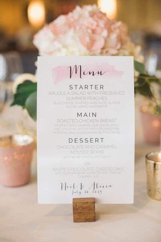 31 steps wedding planning checklist 27 Blush-and-ivory wedding reception setup featuring a modern calligraphy wedding planning checklist card on a wooden stand, surrounded by hydrangeas and votive candles.