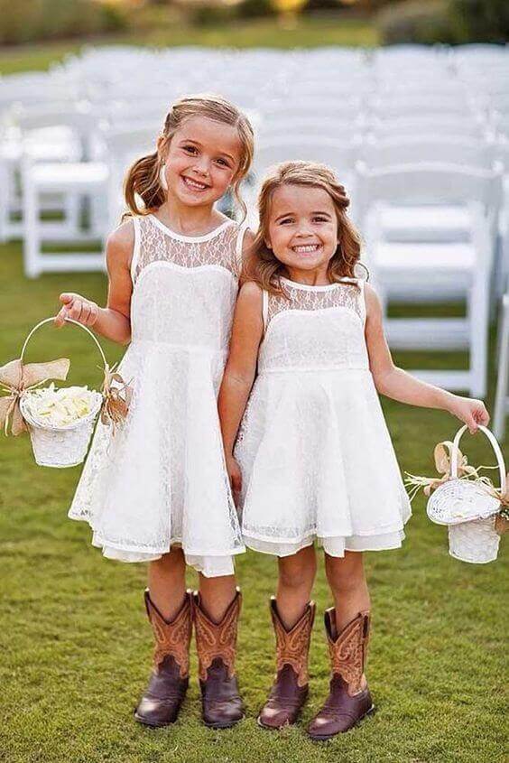 43 cute and perfect flower girl dresses that make every little princess shine 28 Two smiling flower girls in white lace dresses and brown cowboy boots stand on a green lawn, holding wicker baskets filled with rose petals.