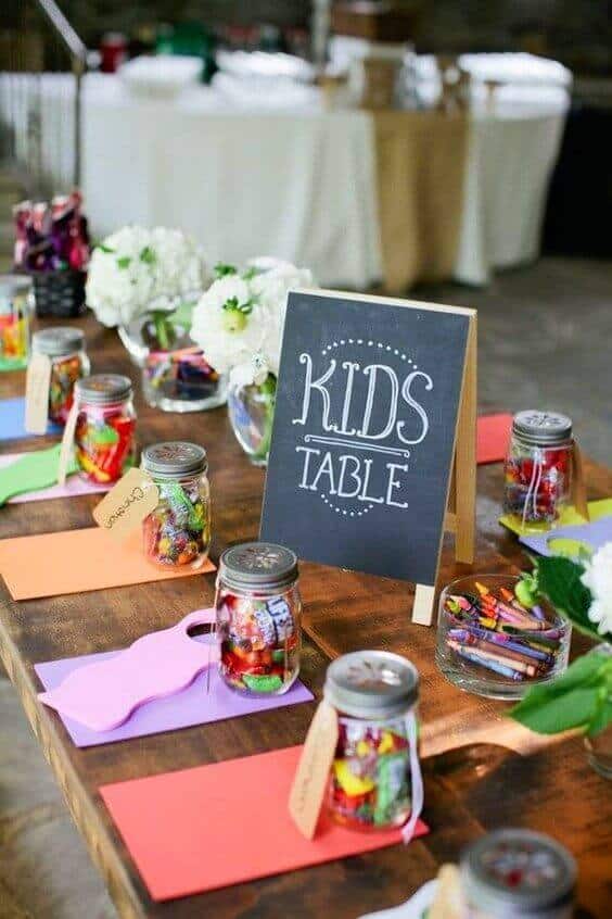 34 budget wedding ideas for a creative affordable wedding 29 A vibrant kids' table at a rustic wedding features a chalkboard sign, mason jar candy favors, colorful placemats, and white floral arrangements.