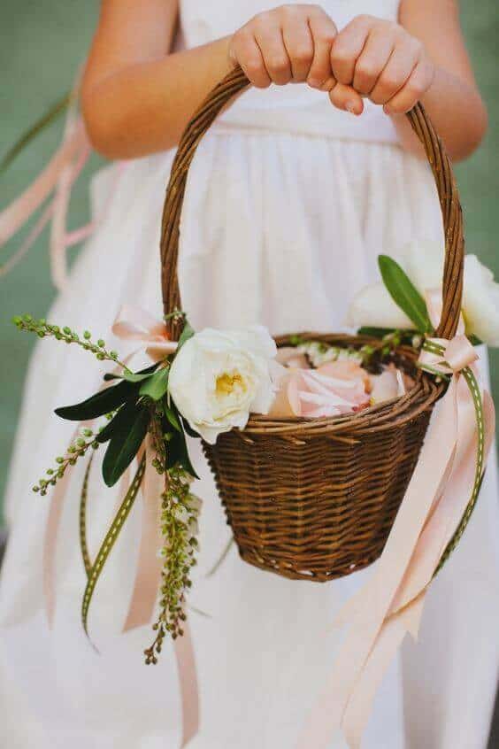 38 wedding accessories list - ideas to transform your big day into magic 29 A close-up of a handwoven wicker flower girl basket filled with blush rose petals and peony blooms, held by a young attendant in a white dress.
