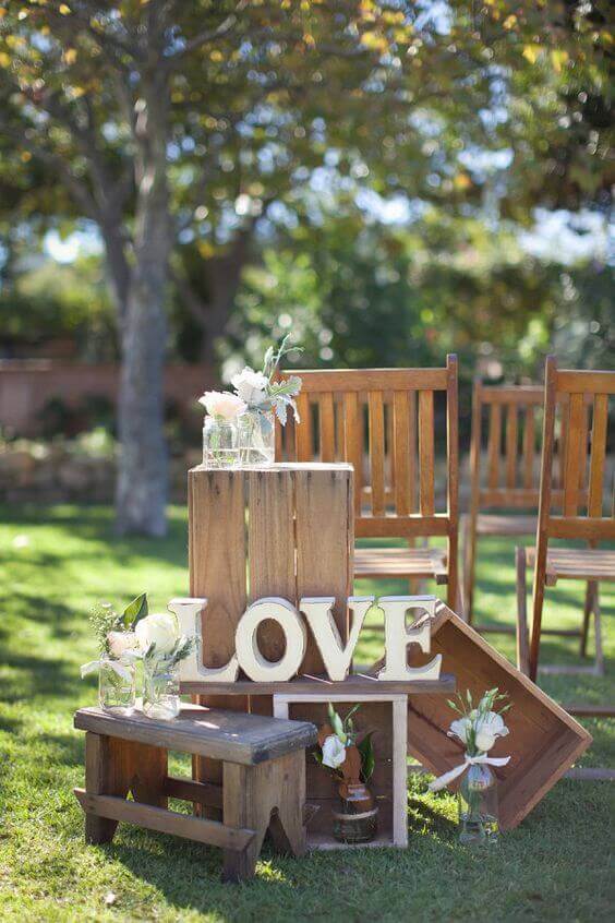 34 budget wedding ideas for a creative affordable wedding 30 Rustic wedding setup featuring stacked wooden crates, "love" letters, mason jars with white flowers, and ceremony chairs under leafy trees.
