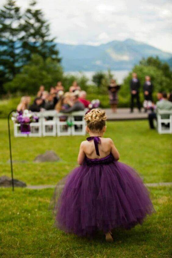 43 cute and perfect flower girl dresses that make every little princess shine 30 A flower girl in a voluminous plum-purple tulle dress with a satin ribbon walks toward a lakeside wedding, framed by mountains.