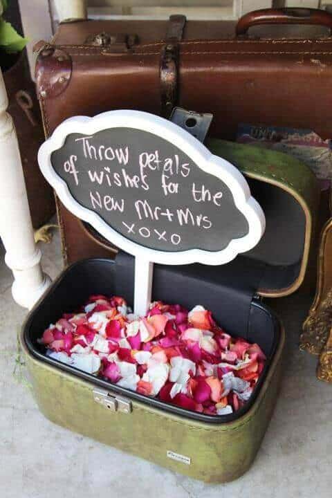 38 wedding accessories list - ideas to transform your big day into magic 30 Open olive-green vintage suitcase filled with rose petals, accompanied by a white cloud chalkboard sign reading, "throw petals of wishes for the new mr + mrs xoxo," styled next to stacked leather luggage.