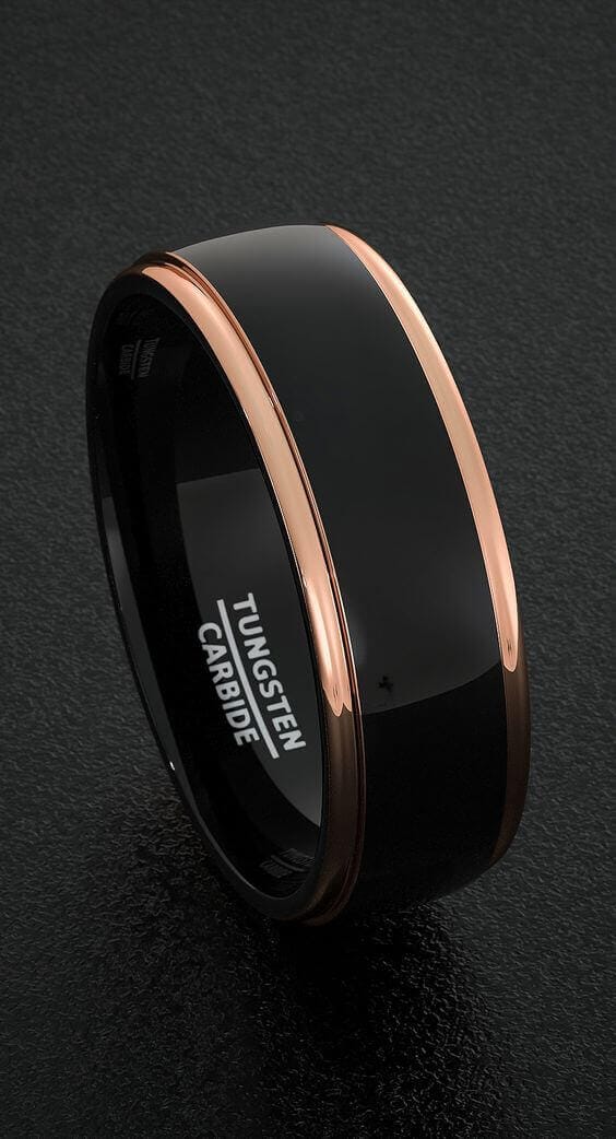 Black tungsten carbide wedding band with glossy center and rose-gold beveled edges, positioned on a textured charcoal surface.