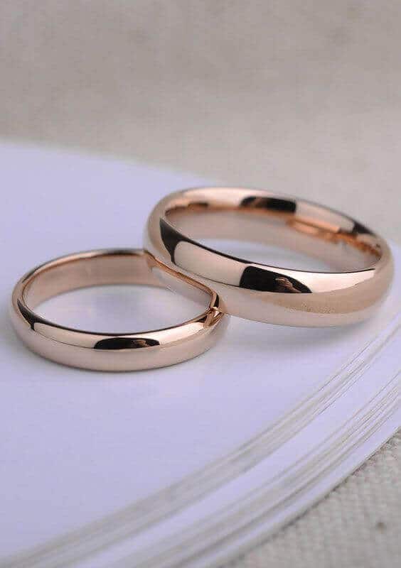 Two mirror-polished rose gold wedding bands gently overlapping on the corner of a white invitation, set against a soft beige linen backdrop.