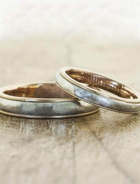 Close-up of two wedding bands on a rustic tabletop; one ring flat, the other overlapping. Artisan hammered finish, mixed metals with satin sheen.