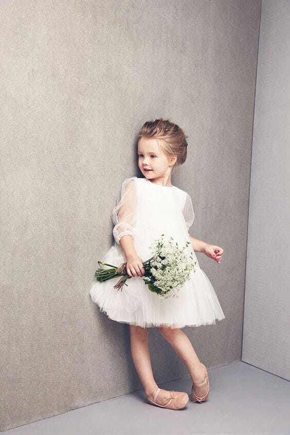 43 cute and perfect flower girl dresses that make every little princess shine 33 Young flower girl in an ivory tulle dress with sheer sleeves, holding a baby's breath bouquet, posed ballet-style against a grey wall.