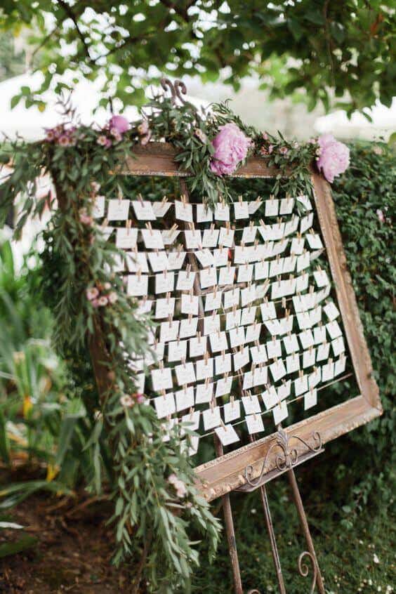 38 wedding accessories list - ideas to transform your big day into magic 33 Rustic wooden frame on a wrought-iron easel displays ivory escort cards, adorned with eucalyptus and blush flowers in a garden setting.