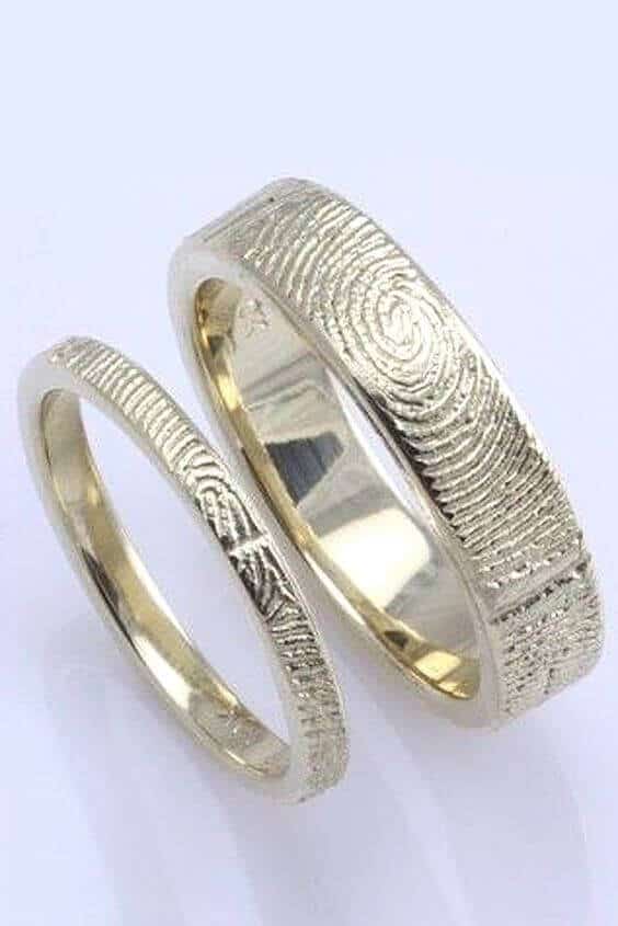 Two yellow-gold wedding bands with fingerprint engravings, one featuring a bold whorl and the other with fine ridges, on a white background.