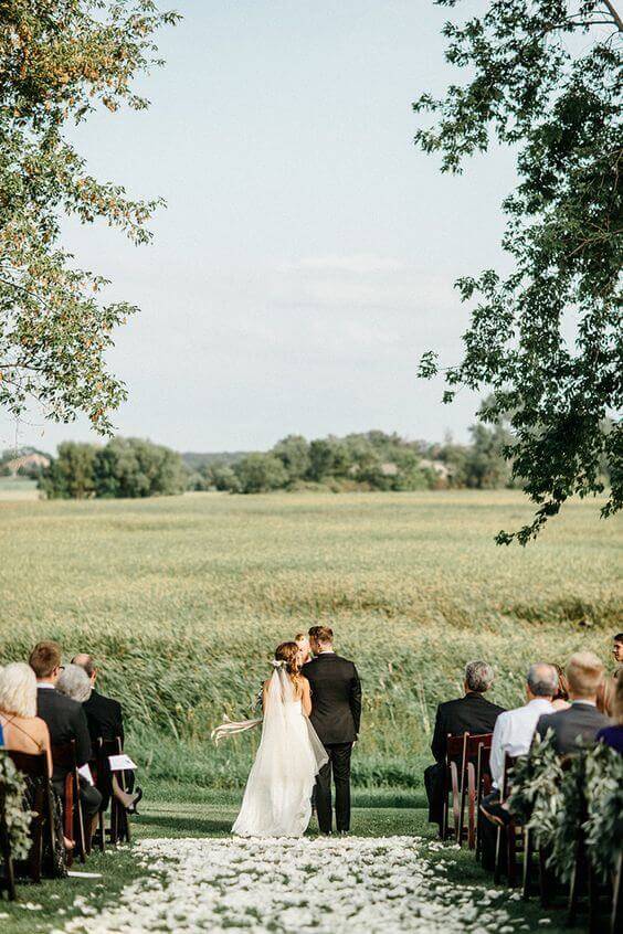 34 budget wedding ideas for a creative affordable wedding 36 An outdoor budget wedding ceremony in a sunlit meadow features a bride in an ivory gown and a groom in a black suit walking down a petal-covered aisle. Dark wood chairs with greenery decorate the rows, framed by two large trees and lush greenery in the background.