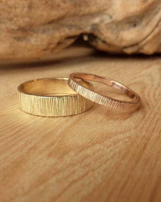 Rustic textured wedding bands on honey-toned wood: a wider matte yellow-gold band and a slimmer rose-gold band entwined, softly lit.