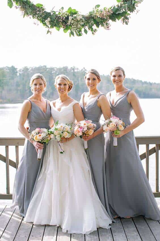 Lakeside portrait of a bride in a white gown with three bridesmaids in dove-gray dresses, holding pastel bouquets, against a serene backdrop.