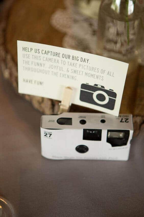 38 wedding accessories list - ideas to transform your big day into magic 39 Rustic wedding reception setup featuring a white disposable camera, a sign for guests, and soft ambient lighting on a neutral table.