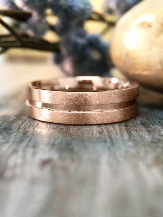 Rose gold double-groove wedding band on weathered blue-gray wood, accentuated by blurred lavender florals and champagne-gold decor.