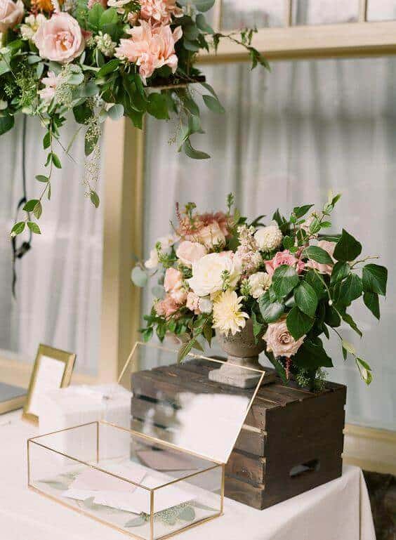 38 wedding accessories list - ideas to transform your big day into magic 40 Blush and ivory floral centerpiece on a wooden crate beside an open glass card box on a white linen wedding welcome table.