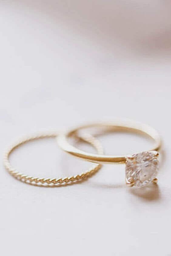 Close-up of a yellow gold solitaire engagement ring with a round diamond next to a thin twisted gold wedding band on an ivory surface.