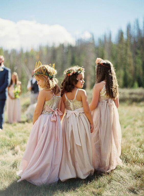 43 cute and perfect flower girl dresses that make every little princess shine 41 Three young flower girls in blush chiffon dresses with gold bodices and oversized satin bows stand in a sunlit meadow, surrounded by greenery.