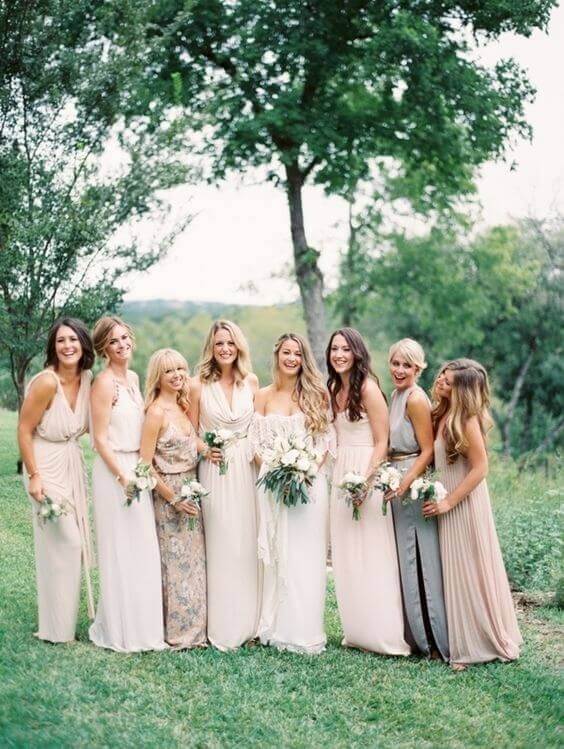 Bridesmaids in neutral mix-and-match dresses stand beside the bride in an ivory gown, holding petite bouquets in a serene garden setting.