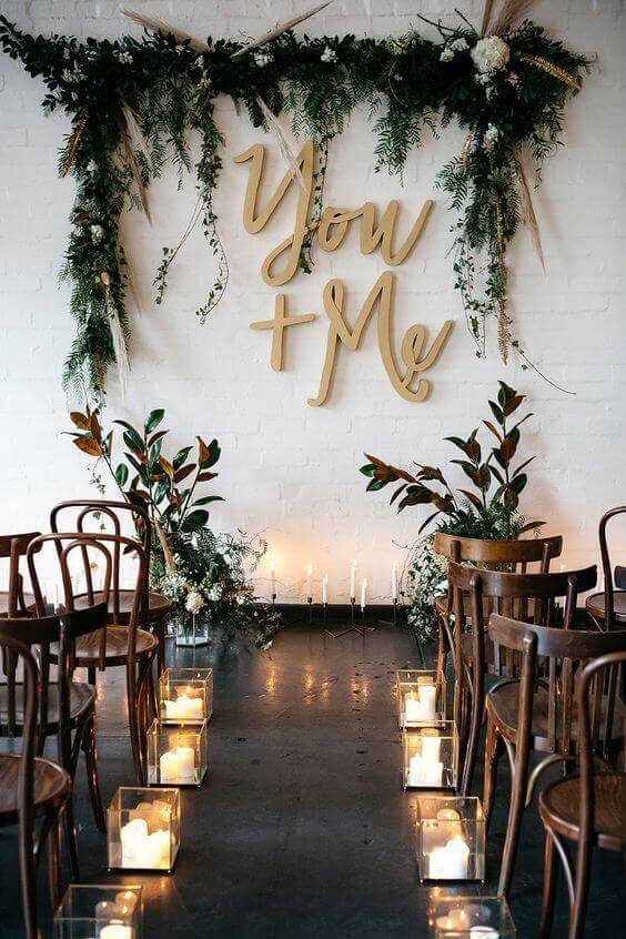 34 budget wedding ideas for a creative affordable wedding 6 Intimate wedding aisle with glass lantern candles, bentwood chairs, greenery garland, and a 'you + me' sign against a white brick wall.